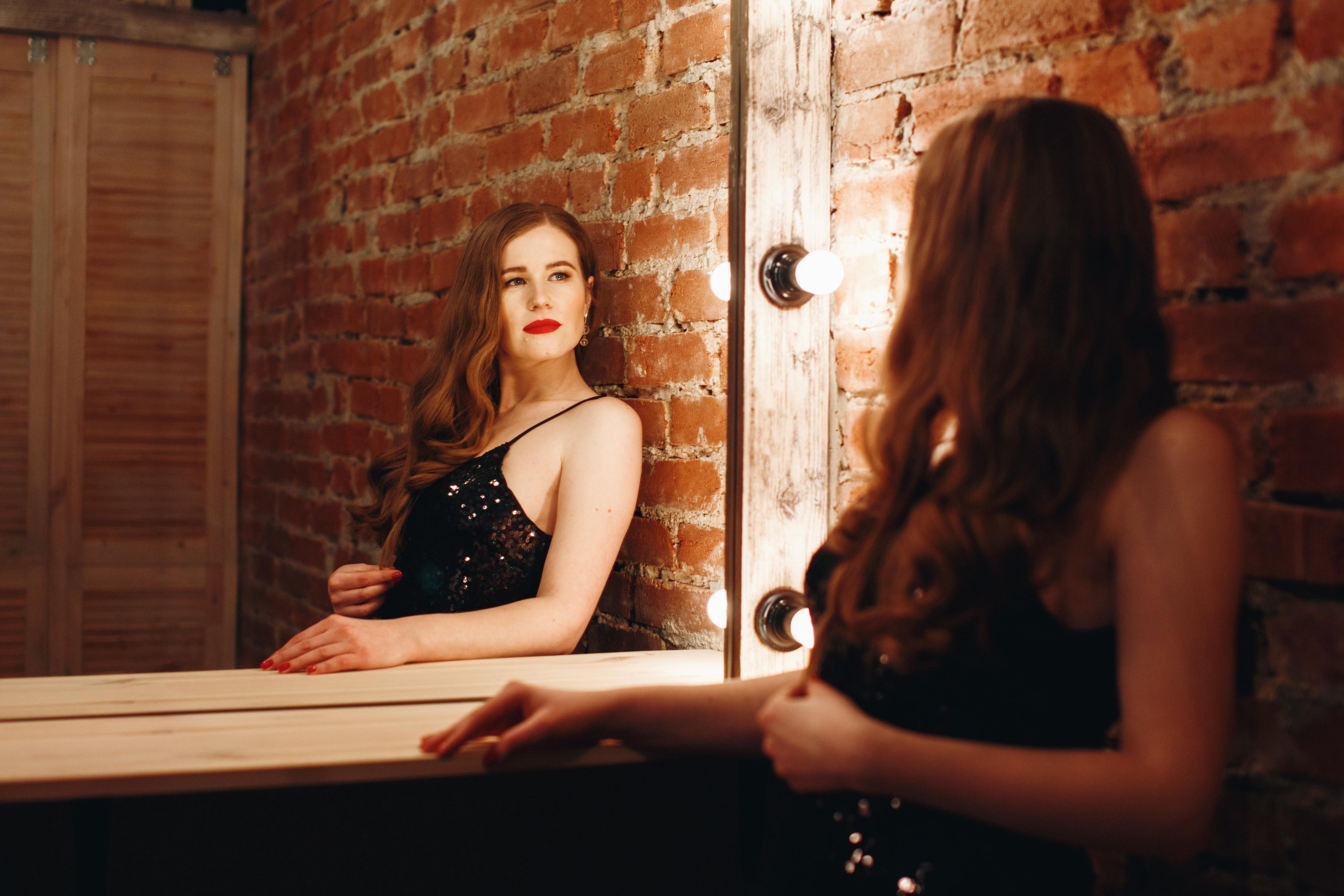 Seductive sexy model posing in loft interior in black dress, looking at vintage mirror with bulbs. Woman with red lips.