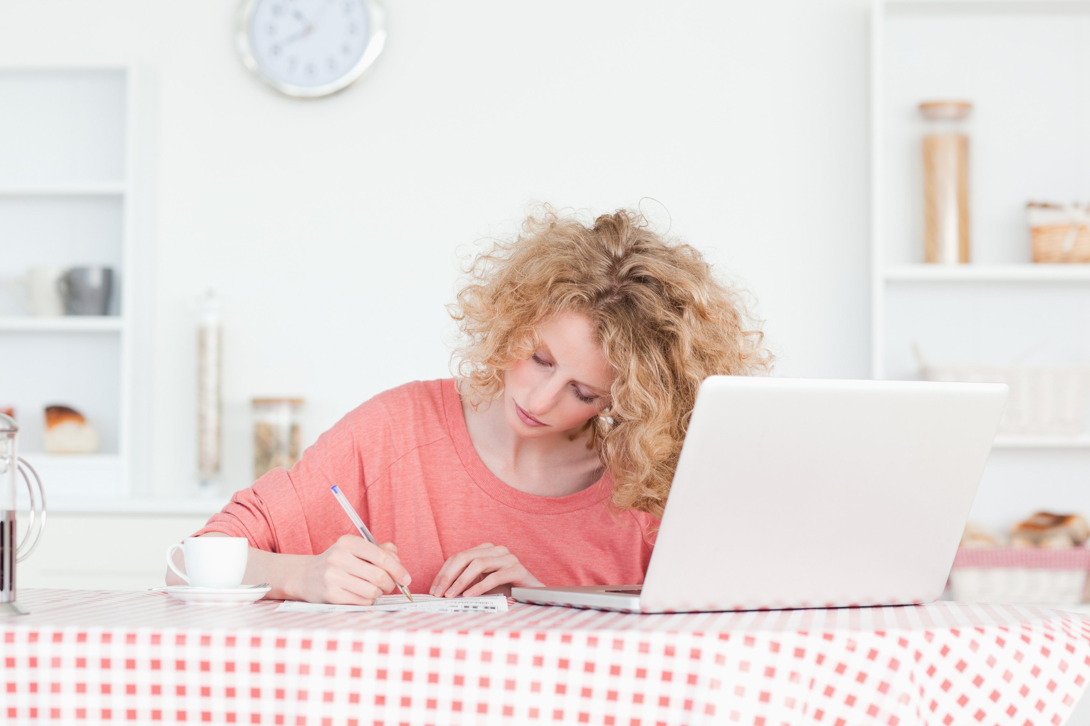 Beautiful blonde woman writing while working with her laptop in the kitchen