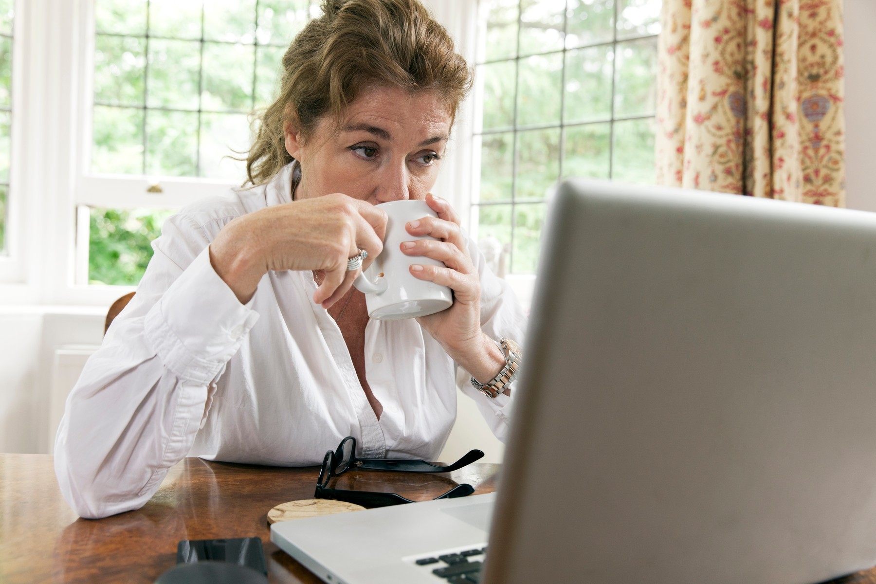 Worried mature woman working on laptop whilst drinking coffee in living room