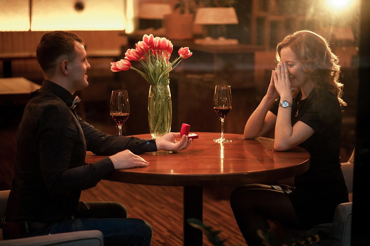 Young man makes woman marriage proposal and offers her jewelry case with engagement ring. They are sitting at table next to glasses of wine and vase w
