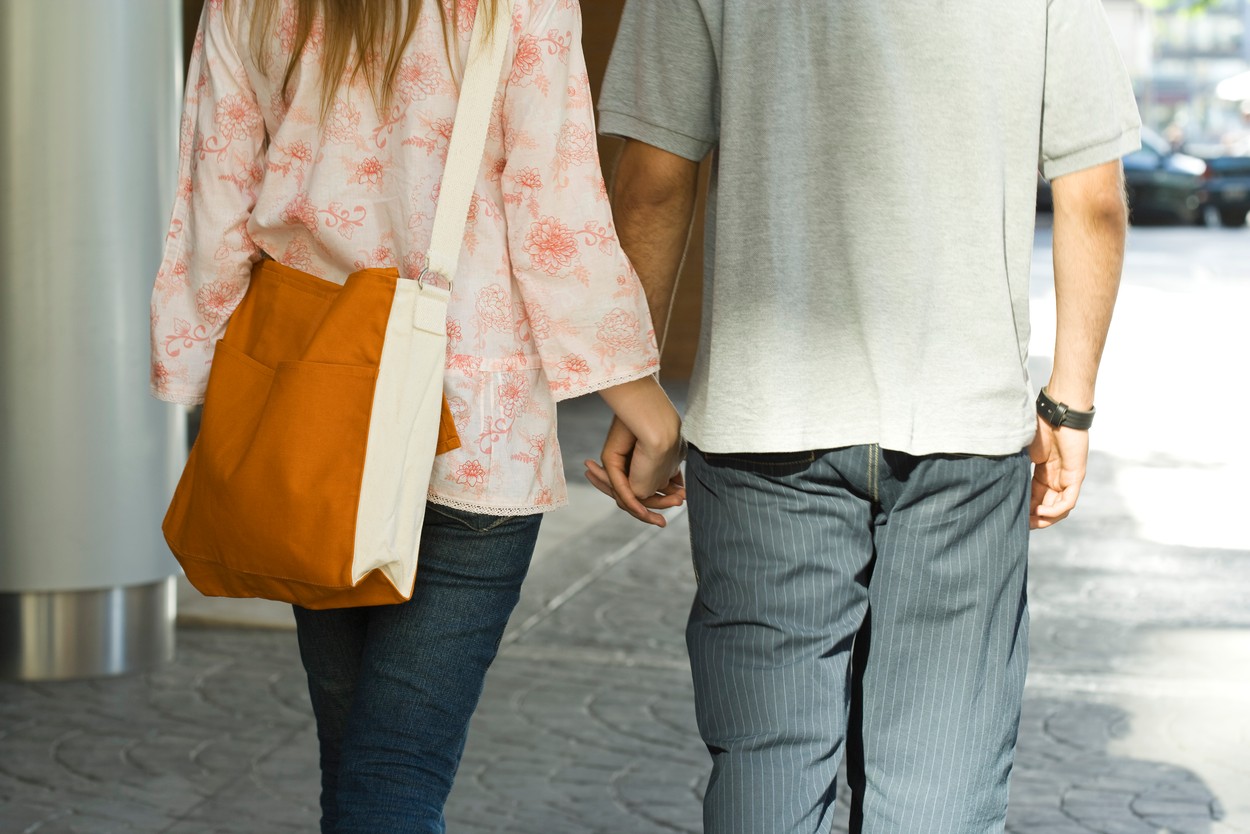 Young couple walking hand in hand on sidewalk, rear view, cropped