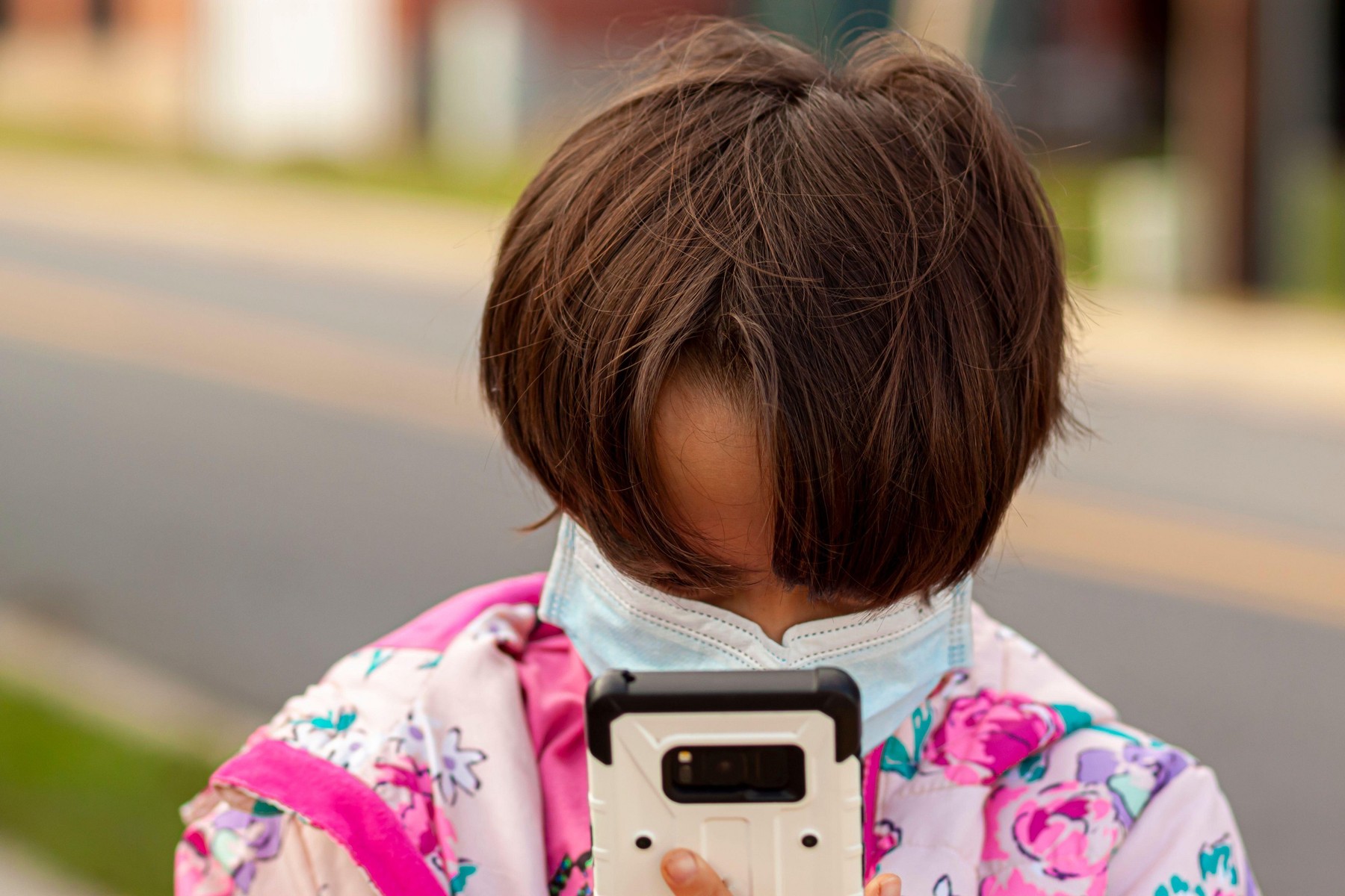 A pretty caucasian girl  with short brown hair, wearing face mask due to COVID-19 pandemic is looking at the screen of a smartphone from close distanc