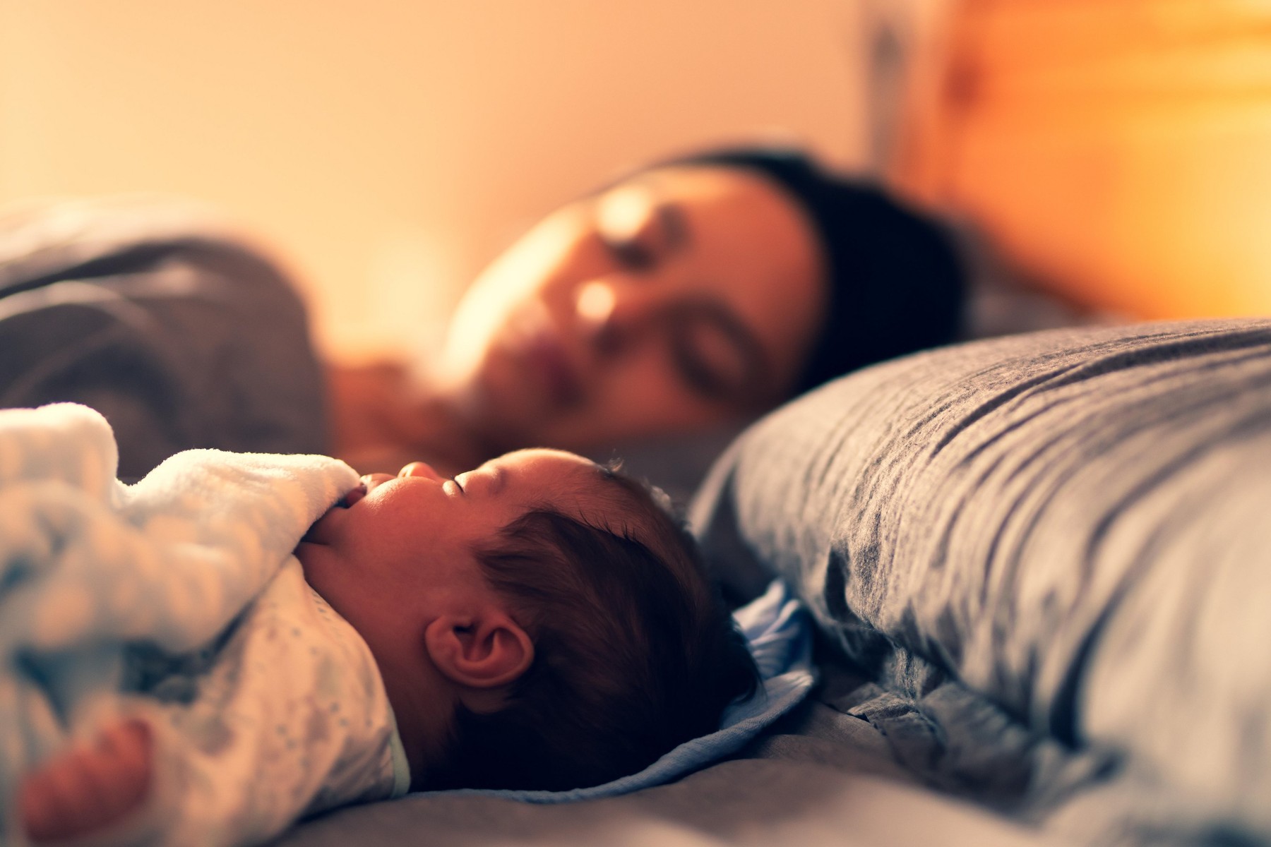 A baby asleep on bed with his mum in the background.