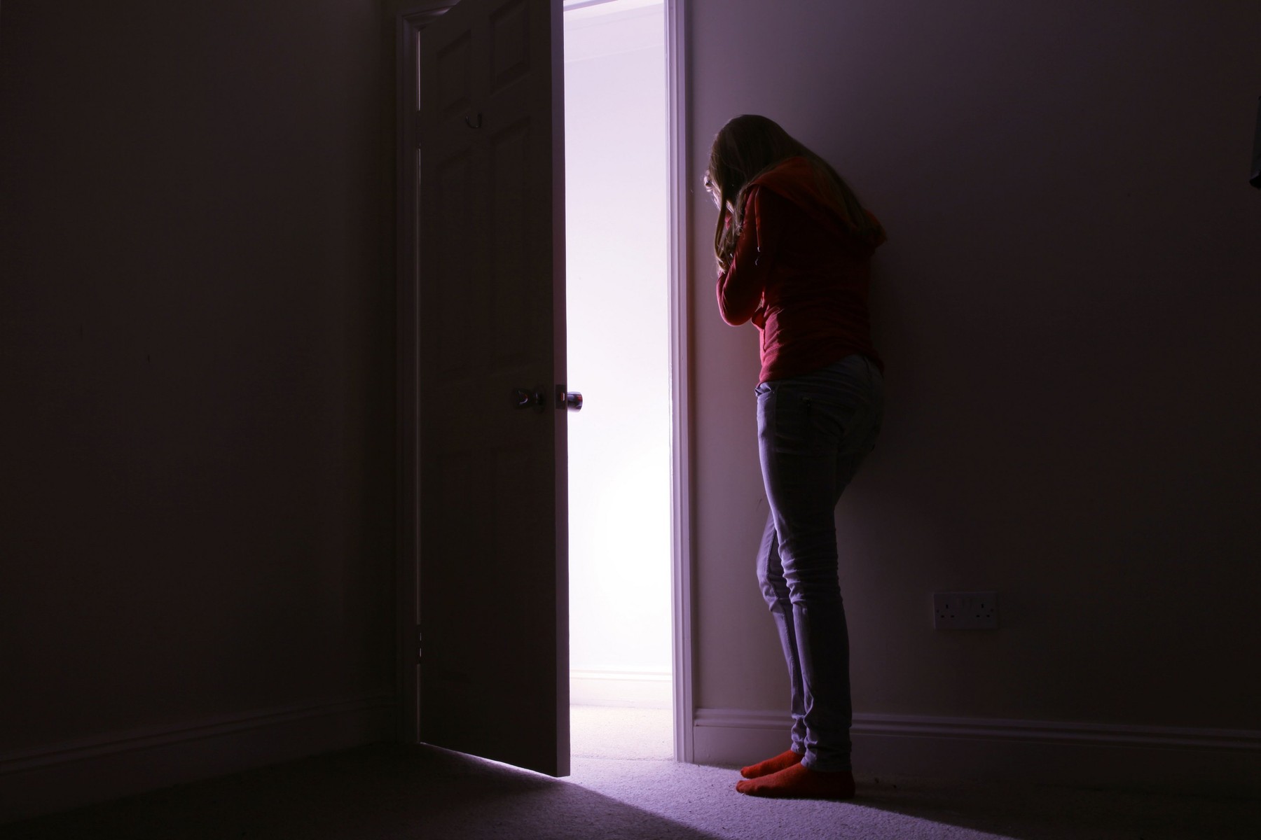 Young female leaning her head against the wall of a dark room, open door with light coming in