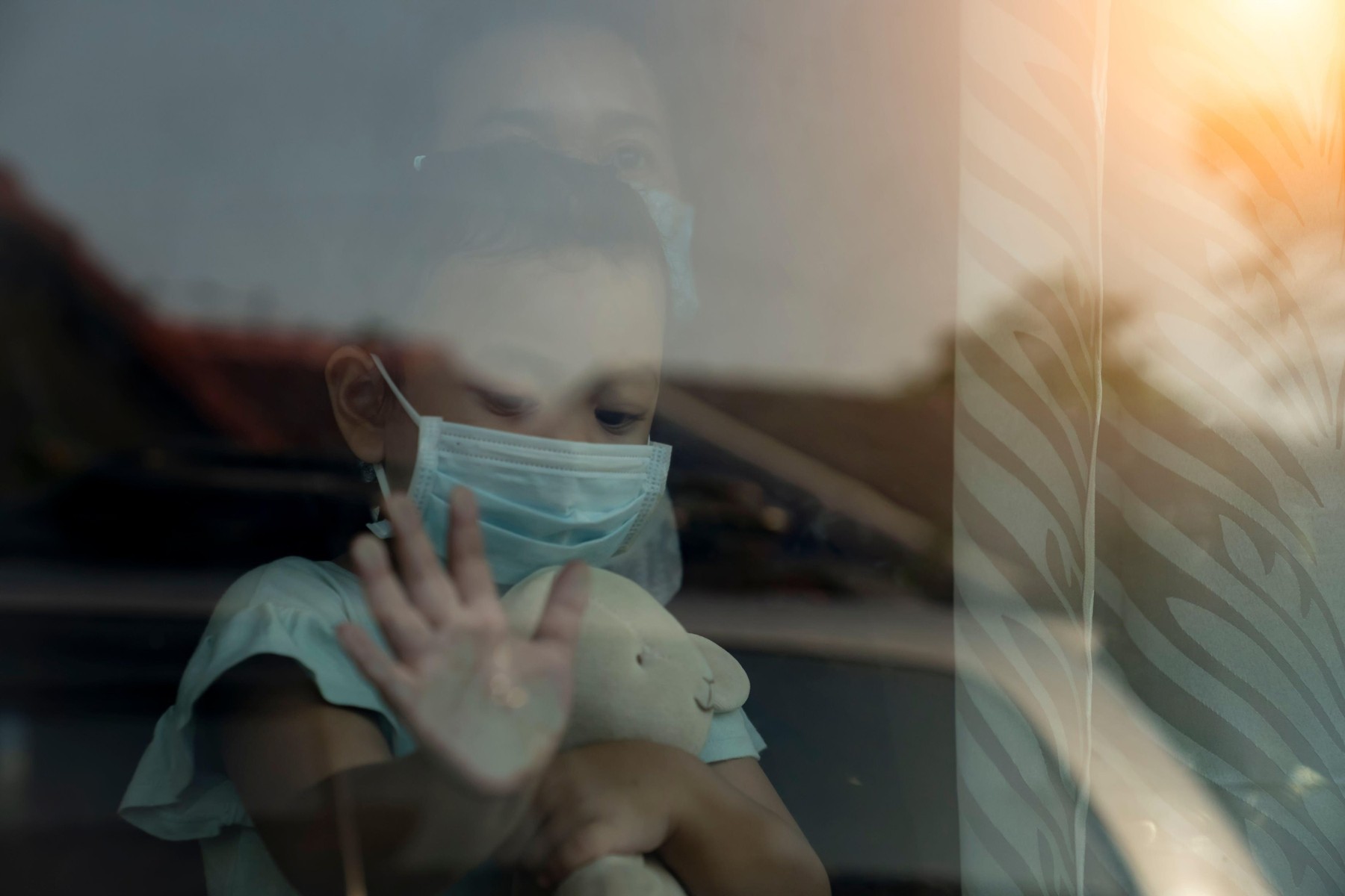 Mother and daughter in protective medical masks standing near a window and looks out window.