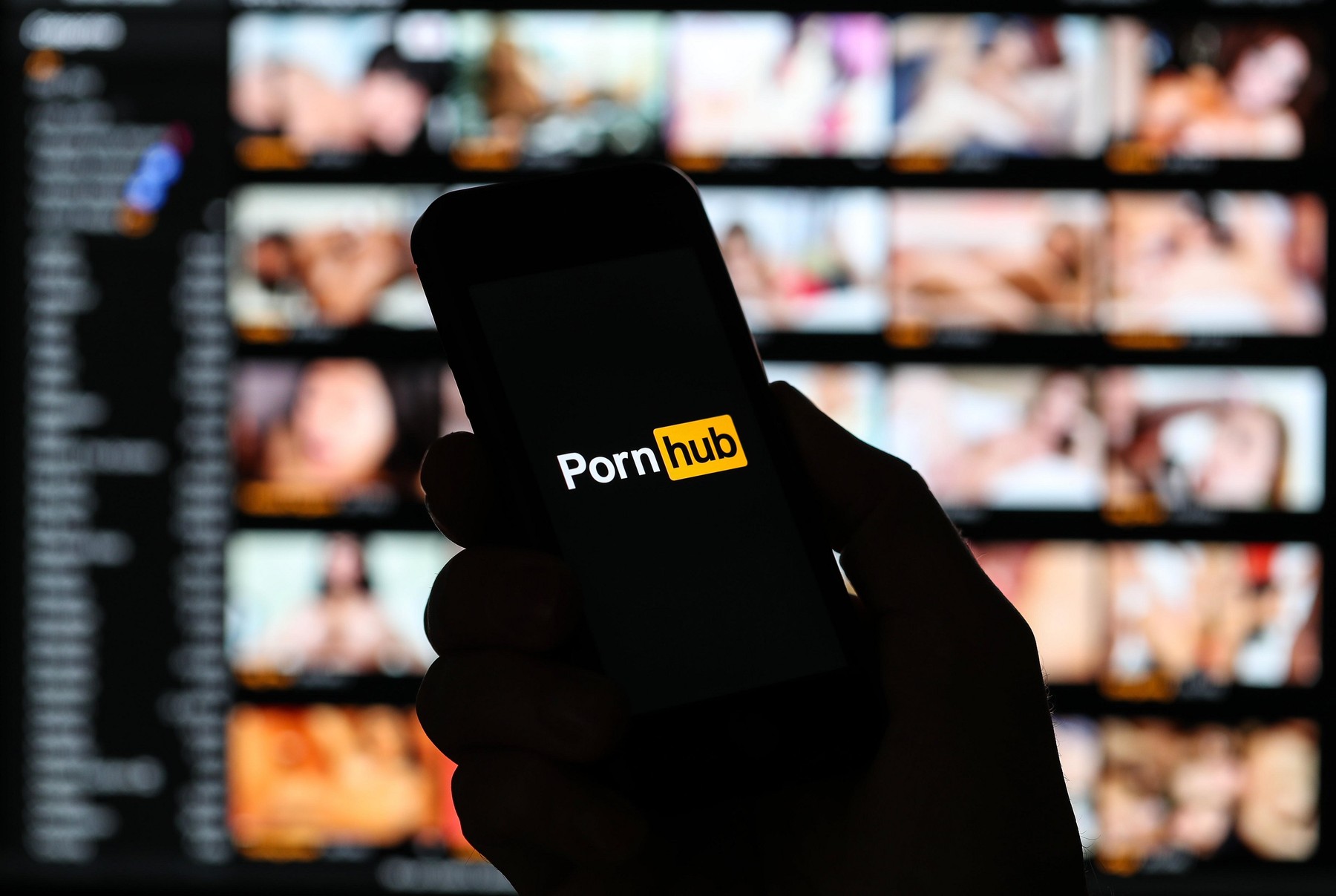 A man looking at the Pornhub.com website logo on his phone with the pornhub website on a computer out of focus in the background.