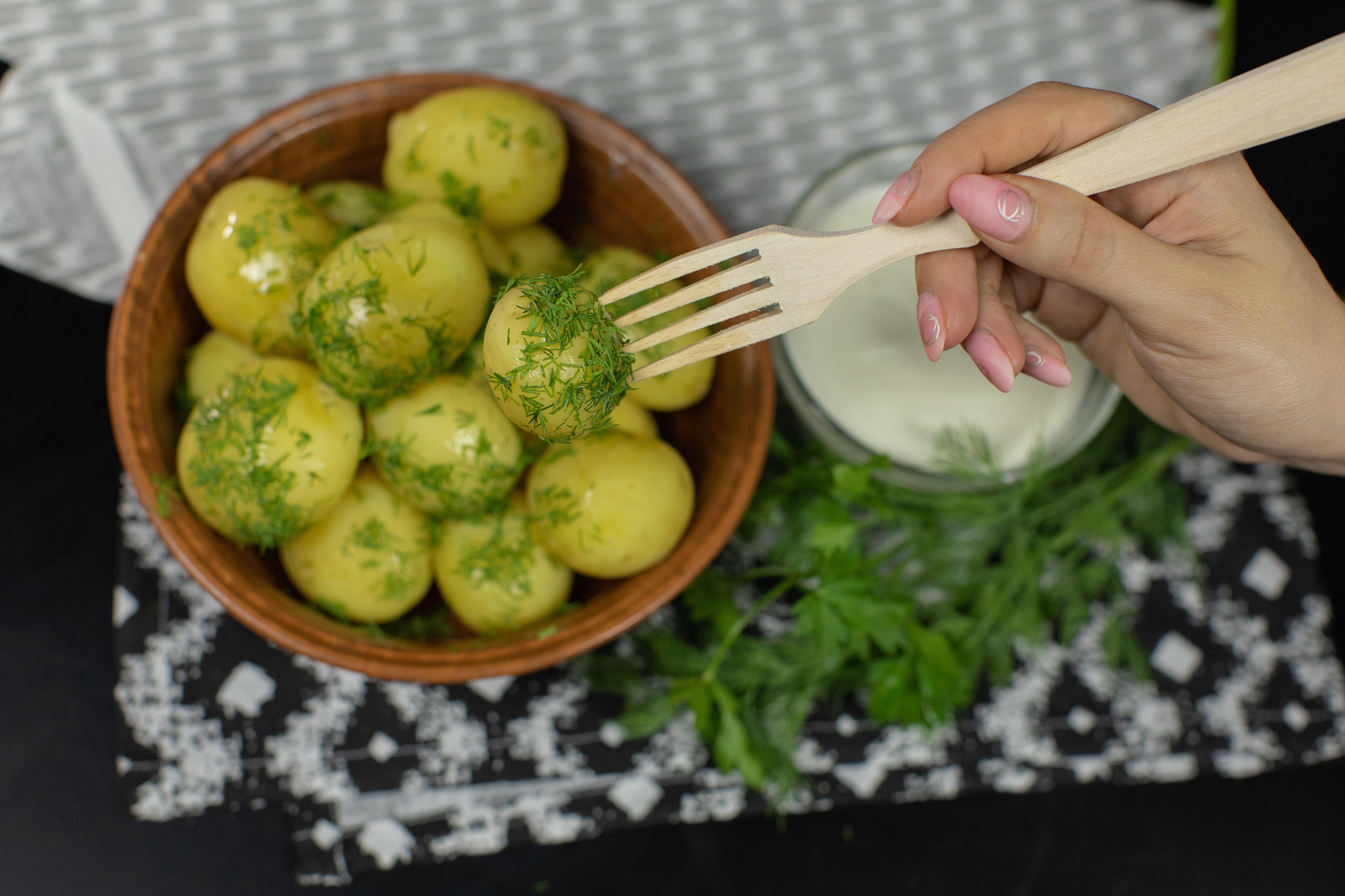 Woman's hand takes boiled new delicious potato using wooden fork from plate on table. Ready to eat. Surface with fresh dill, parsley and sour cream