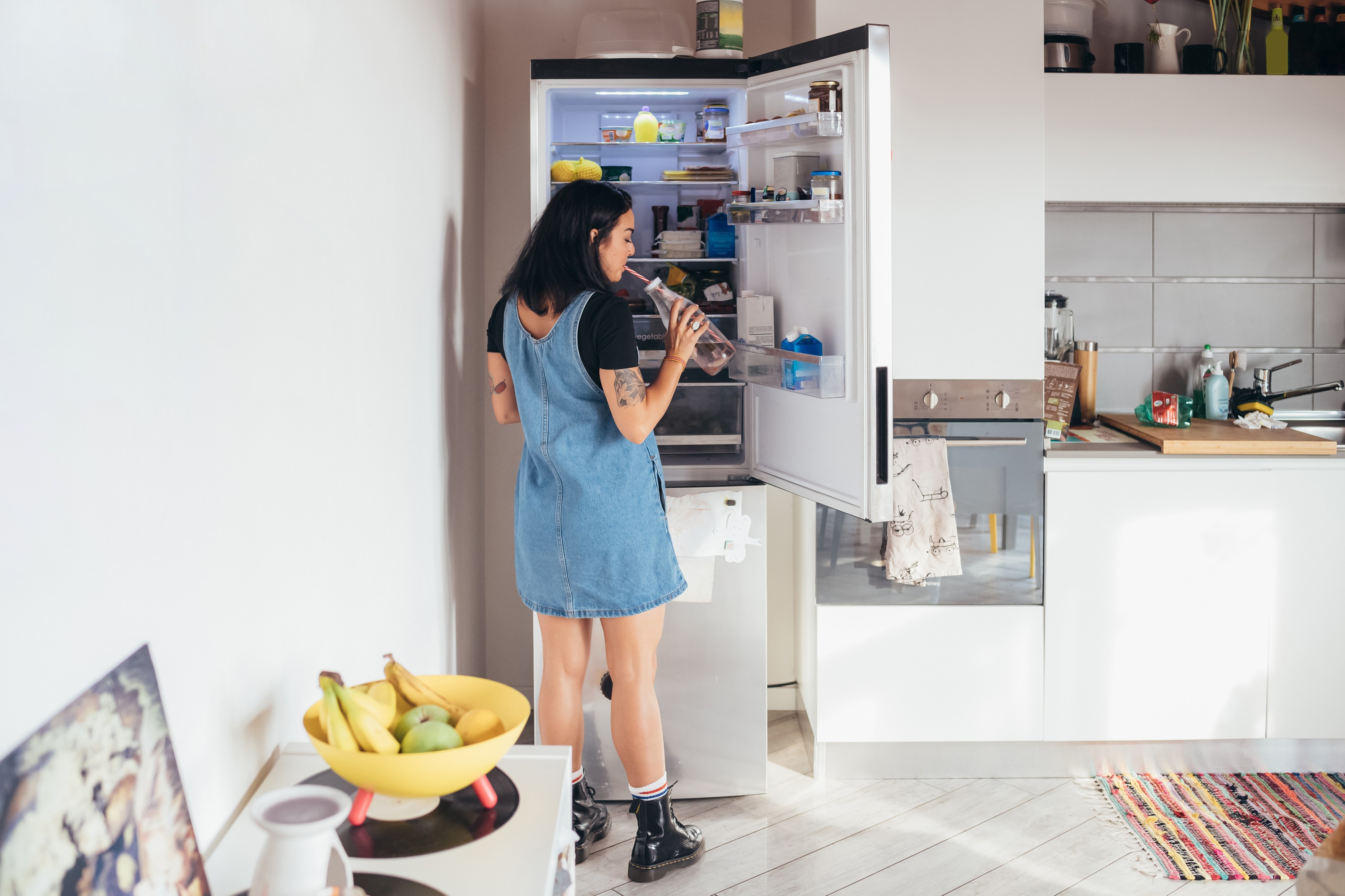 Rear view of tattooed woman with long brown hair wearing denim dress standing in front of open fridge, drinking from bottle.