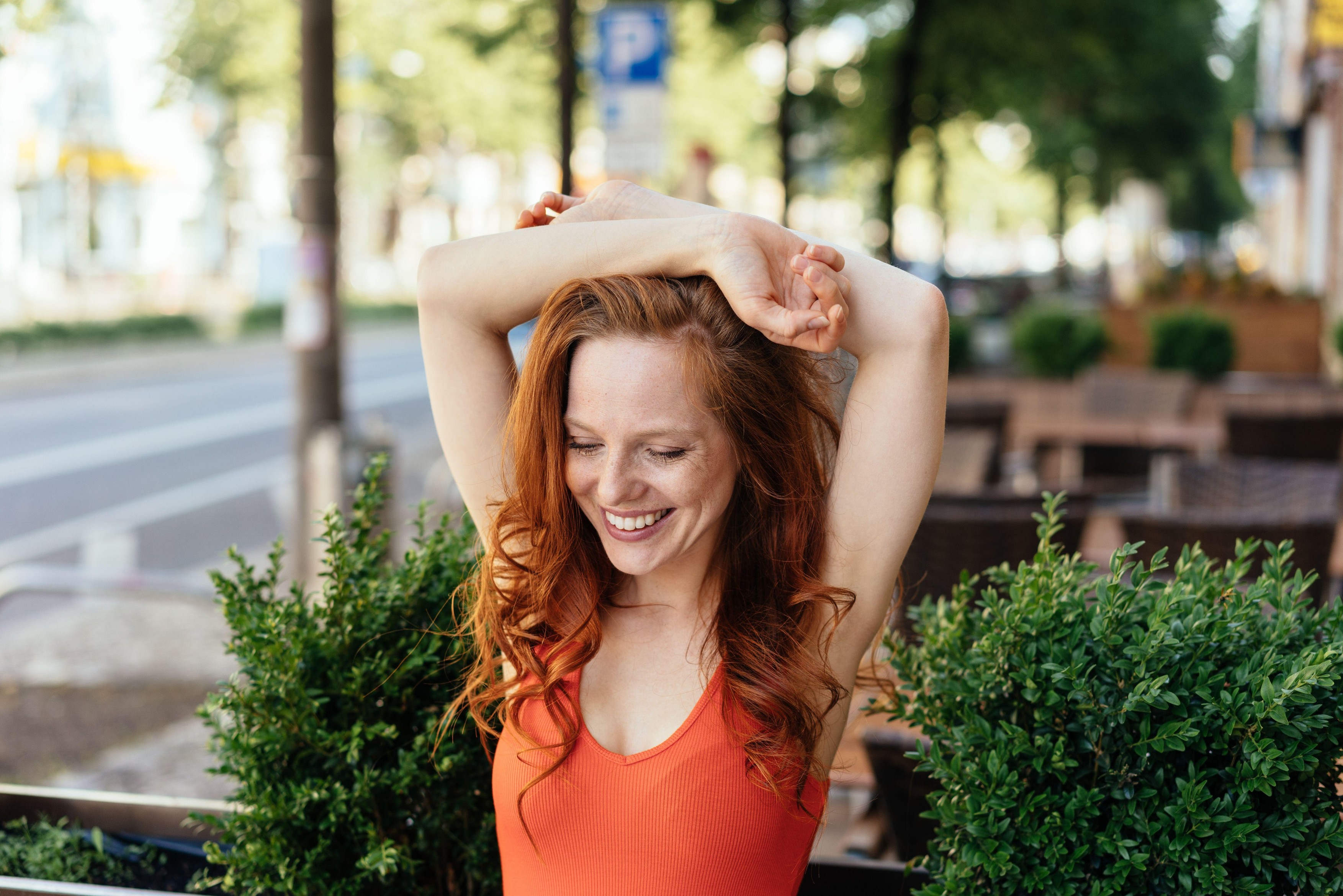 Cute young woman with long curly red hair and a beaming happy smile standing outside a street restaurant in town with arms raised above her head