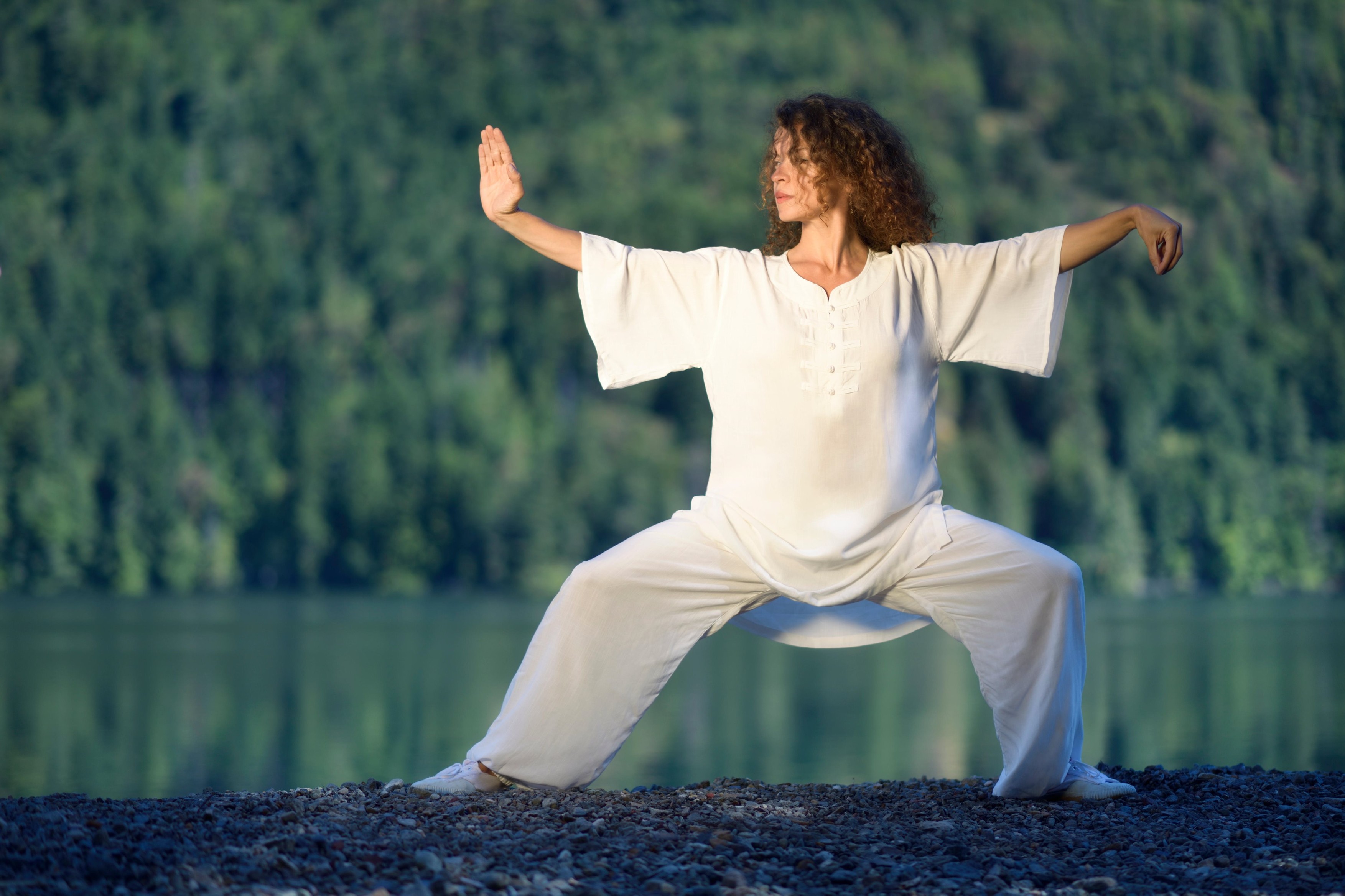 Young woman in white silk uniform practicing a Tai Chi, Taiji, Ban Ma Bu stance in the morning sunrise by the mountain lake in the nature