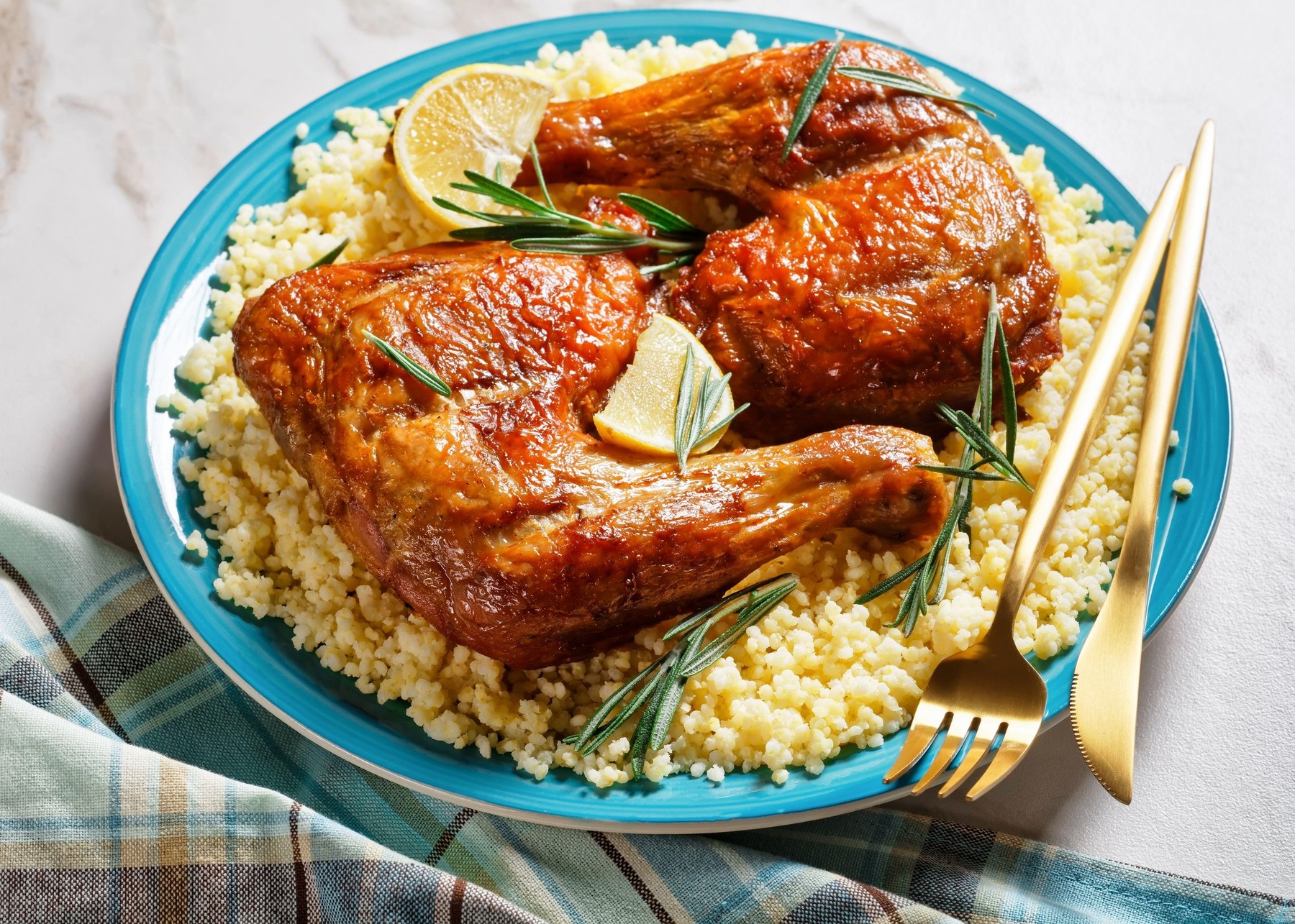 Chicken leg quarters served over the millet on a plate with golden cutlery, lemon wedges, and fresh rosemary sprigs on a white marble stone background