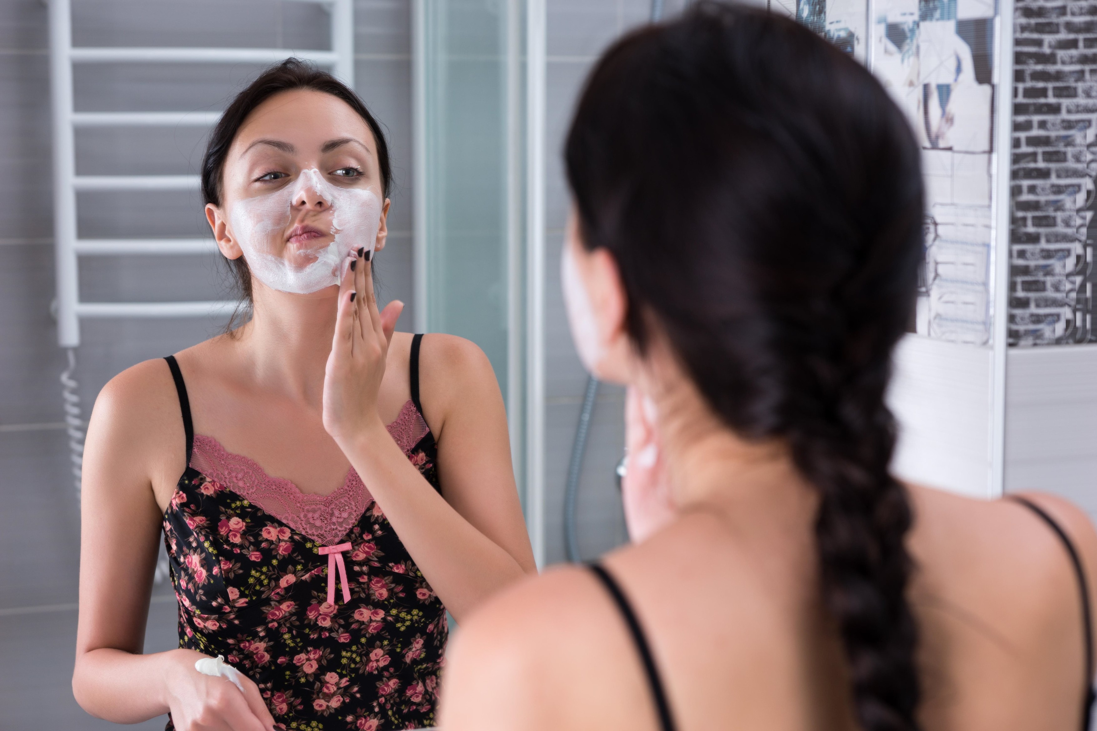 Young woman applying natural facial mask standing in front of  mirror in  the modern tiled bathroom at home