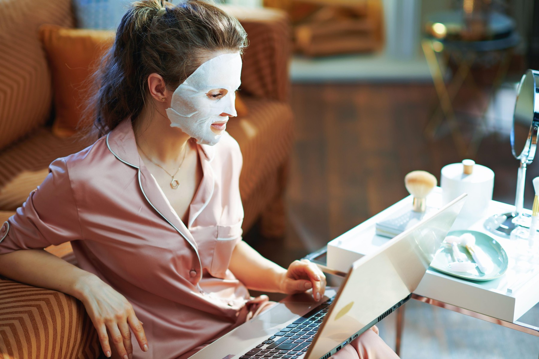 elegant middle age housewife in pajamas with white sheet facial mask near table with toiletries and mirror surfing web on a laptop in the modern livin