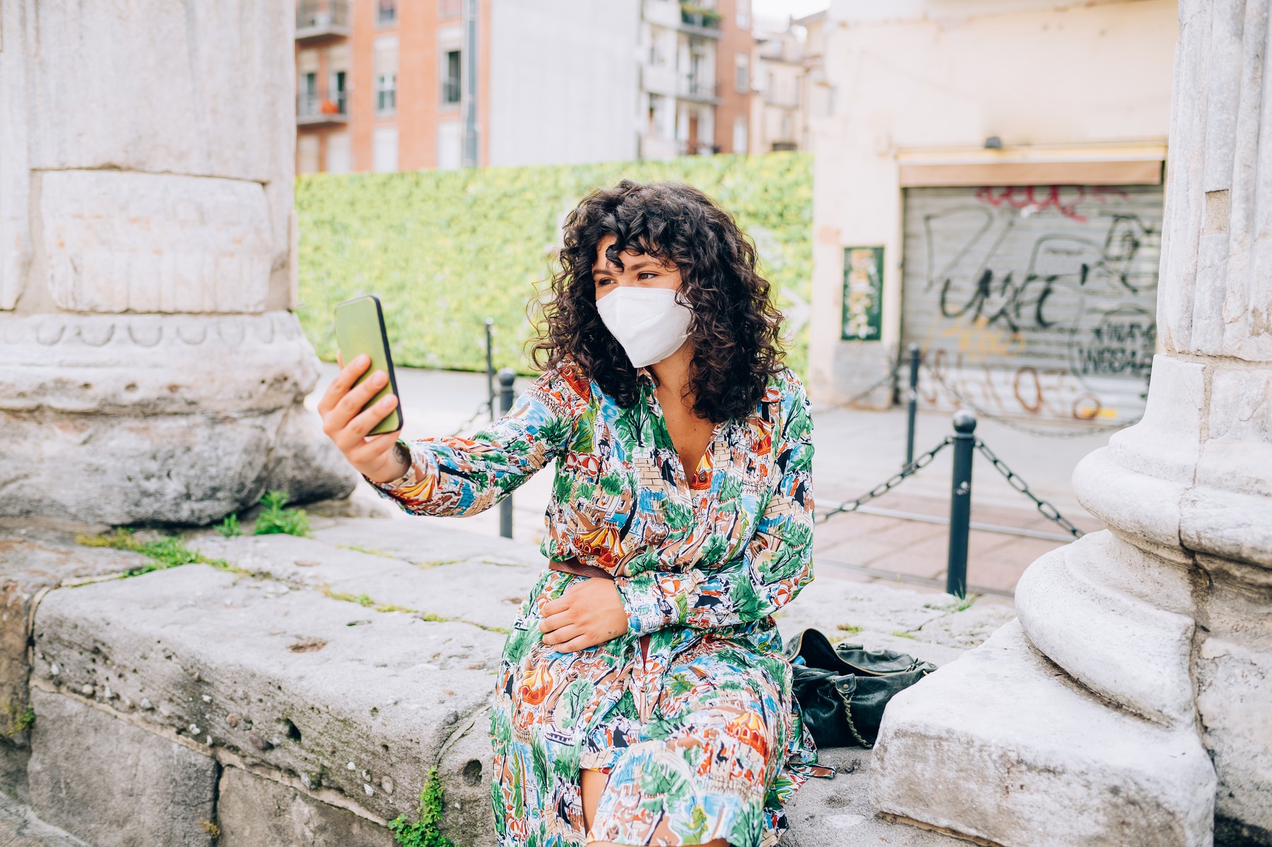 Young woman wearing face mask during Corona virus, sitting outdoors, taking selfie.