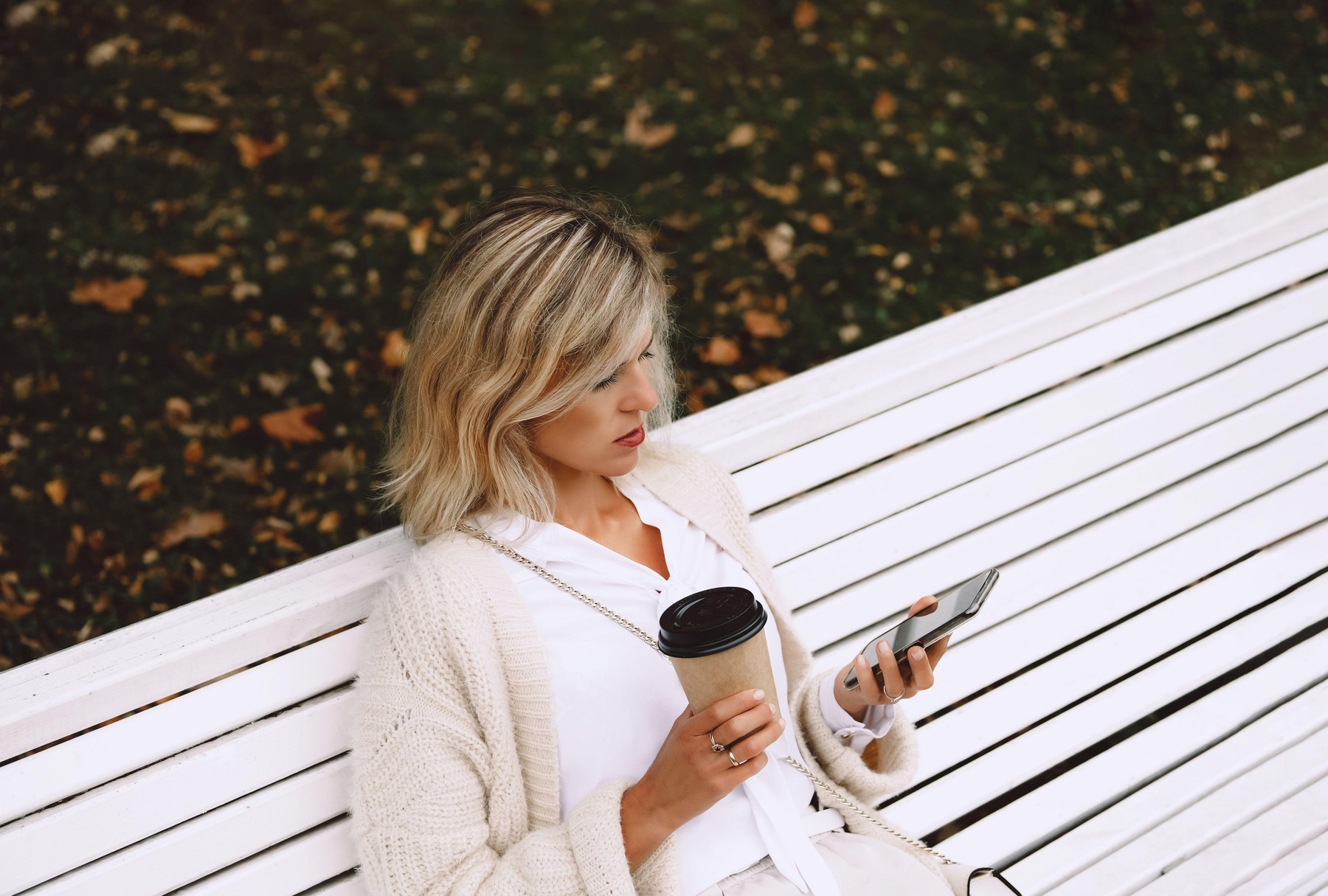 young businesswoman sitting on bench in autumn park and talking over the phone taking notes. Coffee break outdoors.