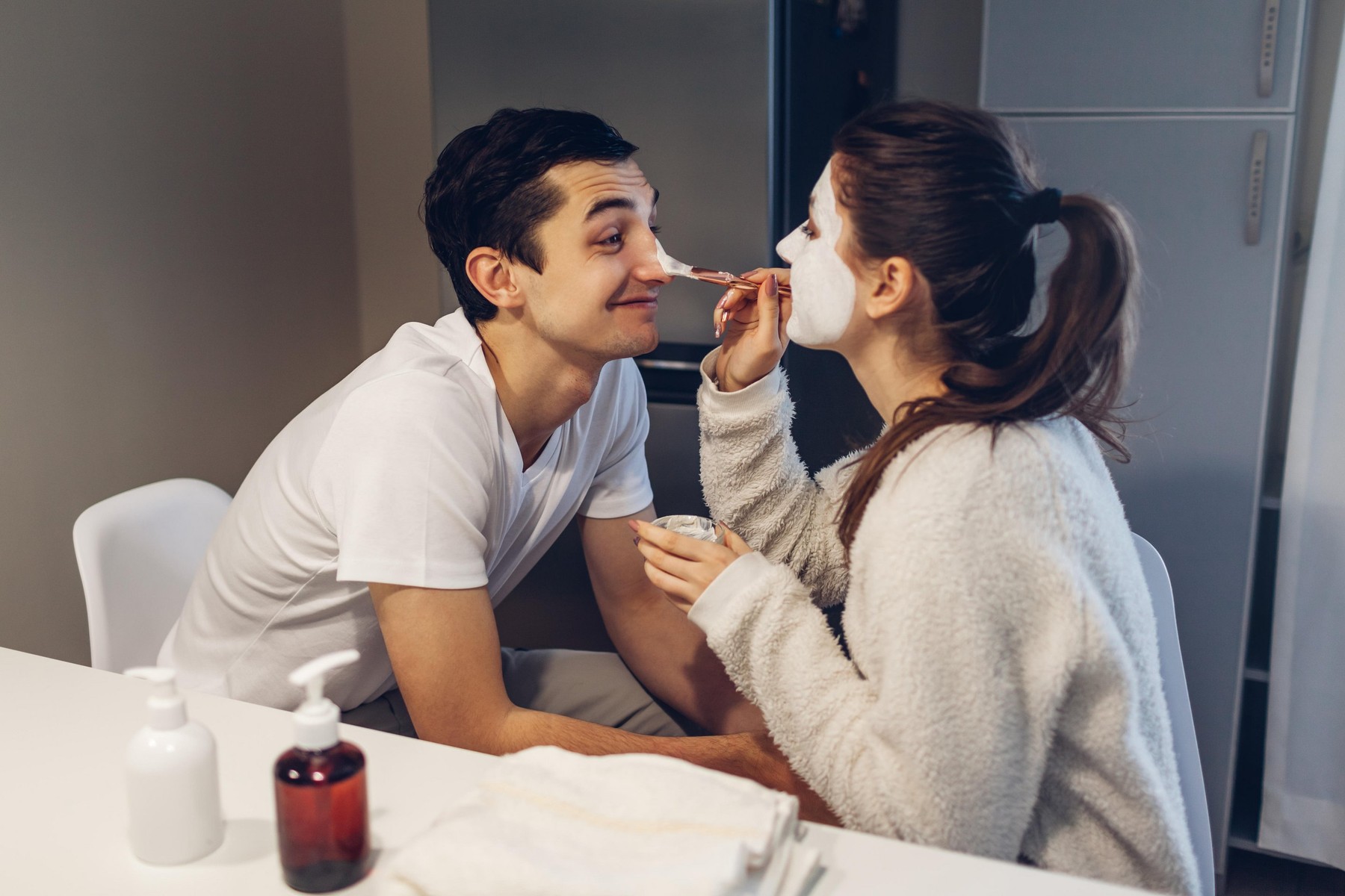 Woman applying clay mask on her boyfriend's face. Young loving couple taking care of skin on kitchen at home