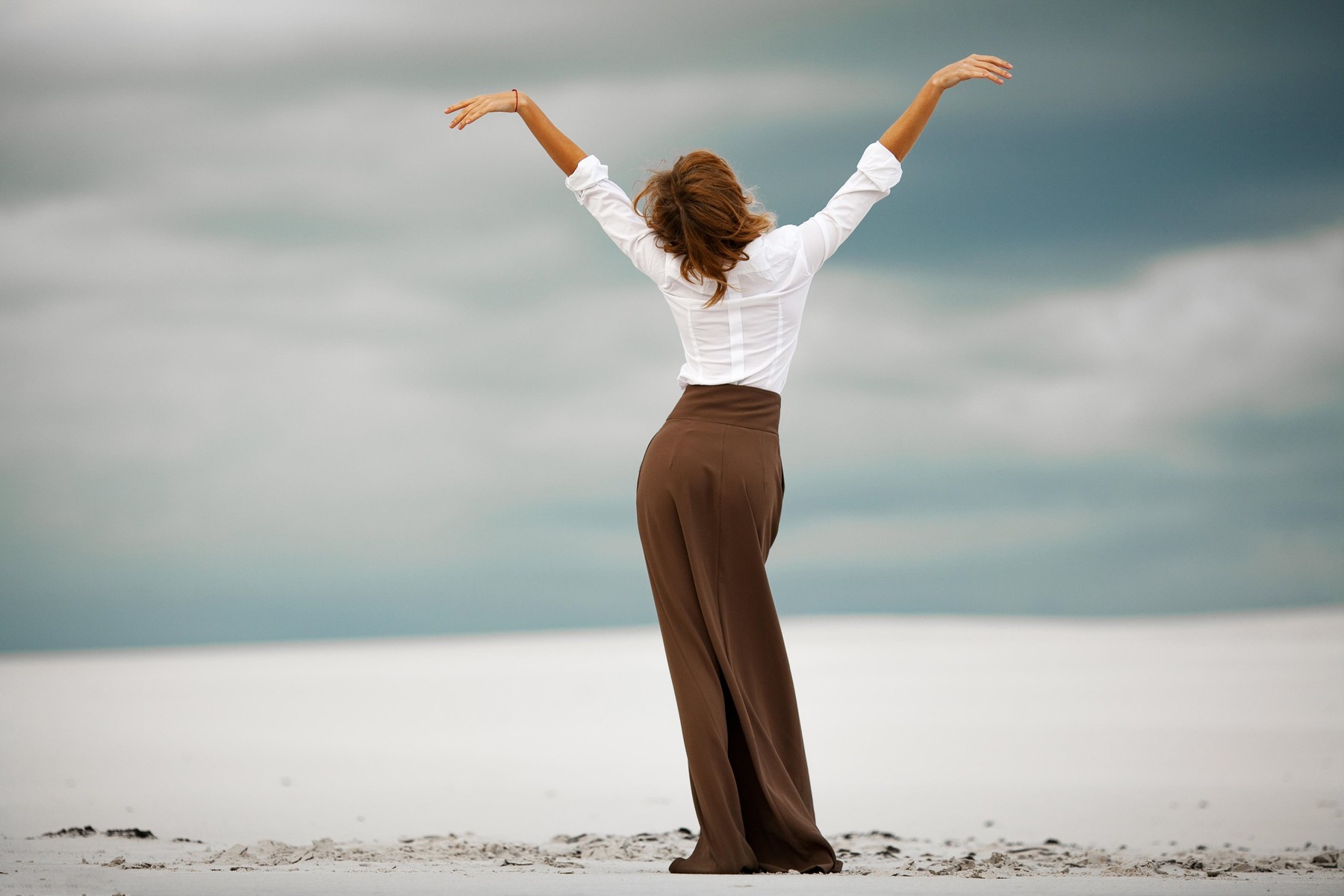 Young woman stands on sand in desert. She is dressed in long skirt and blouse. Back view.